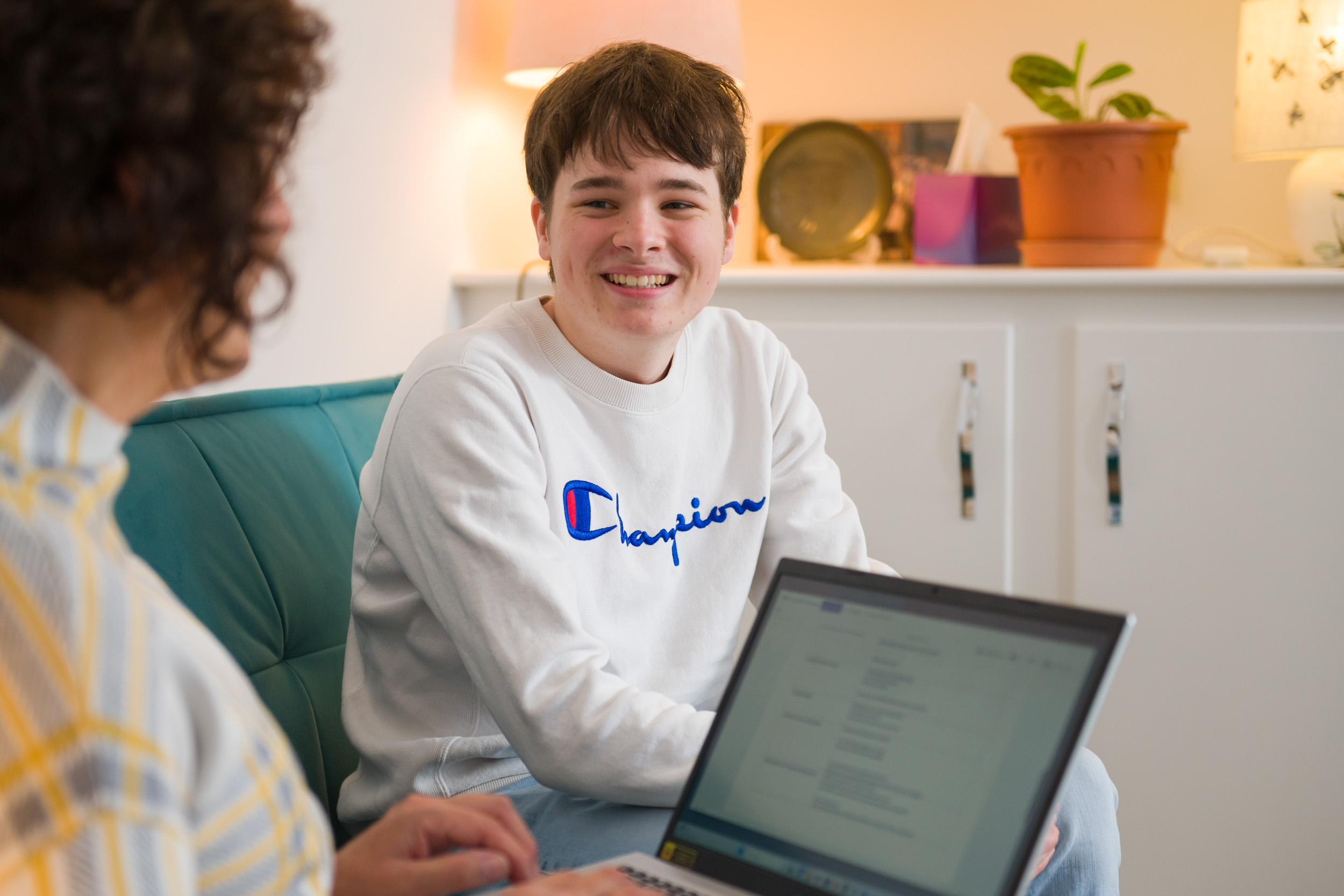 Open Homes Nottingham volunteer with a laptop mentoring a young man on a teal couch in a cosy living room, supporting homeless youth